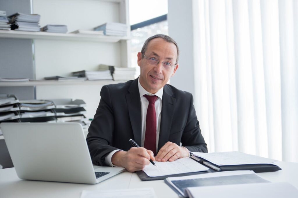 Businessman in a suit writing in an office setting with a laptop and documents.